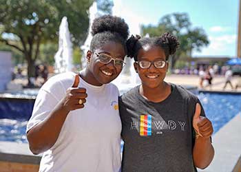 Two Texas A&M university students standing in front of a fountain giving the thumbs up sign with their hands.
