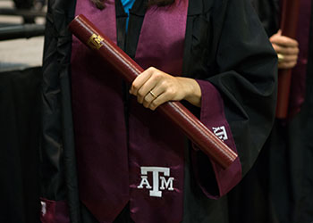 Texas A&M University student wearing graduation regalia holding a diploma tube at the graduation ceremony.